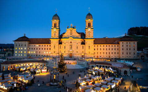 Weihnachtsmarkt Einsiedeln bei Nacht: Der Weihnachtsmarkt in Einsiedeln erstrahlt im Lichterglanz mit beleuchteten Ständen und einer festlich geschmückten Klosterkirche im Hintergrund.